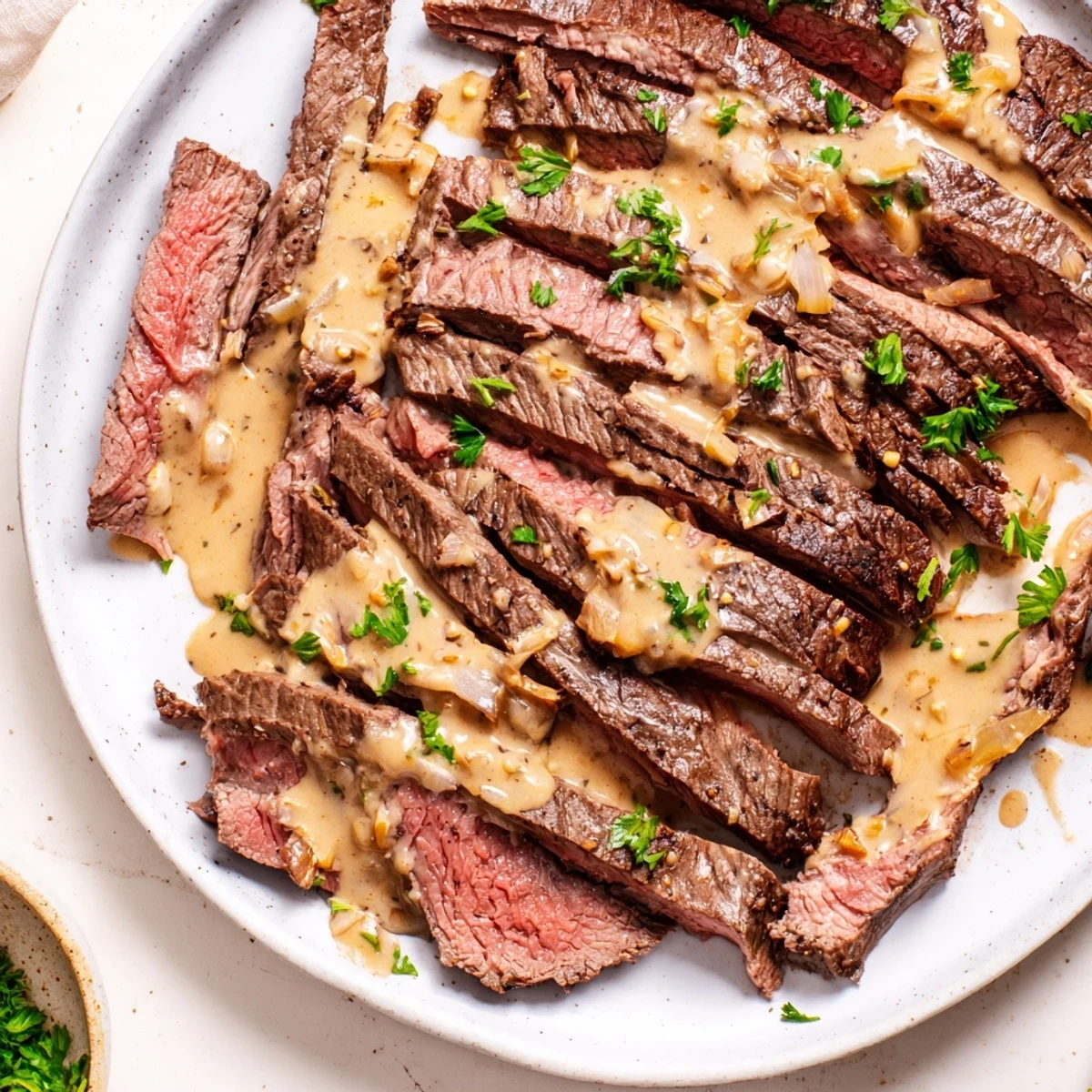 Sliced flank steak with roasted garlic sauce drizzled over, garnished with fresh parsley on a white plate.