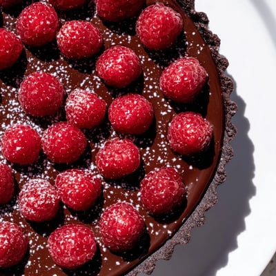 A close-up view of a Chocolate Raspberry Tart slice, showing the crisp chocolate crust and silky ganache layered with ripe berries.